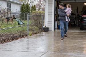 A man carries a child away from a fenced yard where a dog stands.