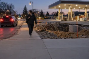 A pedestrian walks beside an open construction trench near a busy road and gas station.