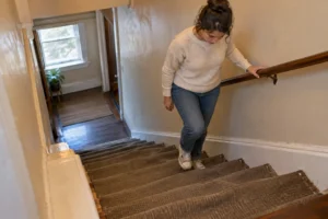 A woman grips the handrail while climbing a narrow, worn staircase in a rental home.