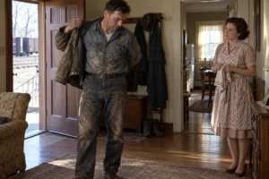 A dust-covered worker removes his jacket while a woman watches inside their home.